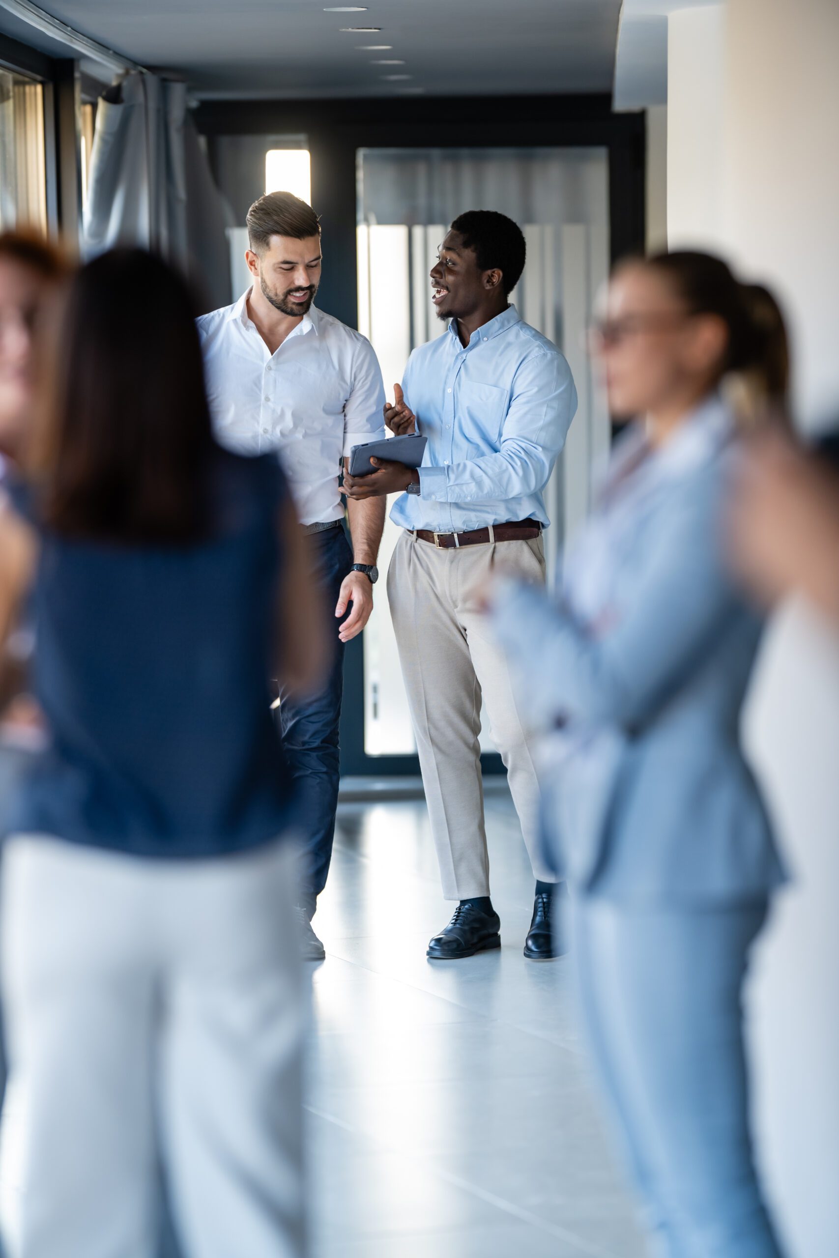 Two businessmen engaged in discussion while colleagues socialize in modern office hallway